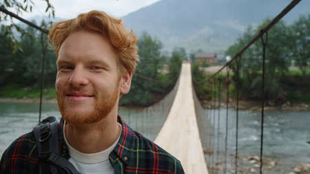 Smiling hiker posing camera on river bridge. Portrait of happy traveler wear backpack on hike. Joyful man enjoy tourism leisure in mountains nature. Ginger guy look around outdoors. Travel concept.の写真素材