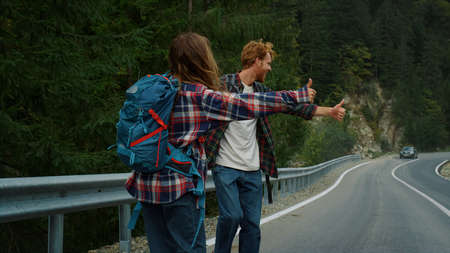 Two backpackers hitchhiking road highway. Couple tourists catch wait car in mountains. Smiling...