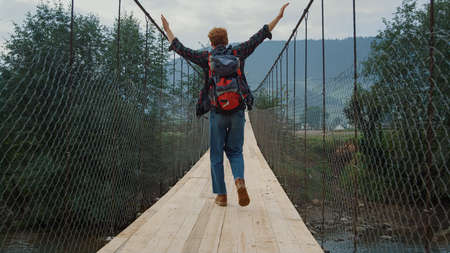 Happy hiker enjoy traveling in mountains forest. Joyful tourist jump on river bridge. Backpacking guy raise hands outdoors. Excited traveler feeling freedom happiness in summer nature. Leisure conceptの写真素材