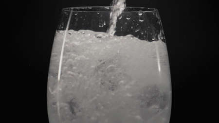 Foaming water pouring ice wineglass closeup. Jet of refreshing bubbled liquid filling transparent...