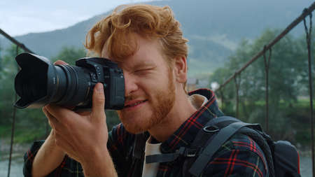 Traveling tourist photographing nature on bridge. Portrait of redhead blogger take photo outside. Relaxed millennial man make picture using digital camera on mountains hike. Holiday summer concept.の写真素材