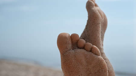 Bare woman feet smeared with beach sand close up. Unknown barefoot girl raising up slim legs sunbathing on sunny seashore. Unrecognizable young lady relax in front beautiful blue ocean summer day.の写真素材