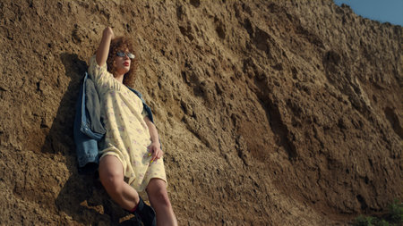 Attractive curly model posing leaning on sand beach hill wearing sunglasses. Stylish gorgeous woman standing seashore in jeans jacket. Fashionable glamour girl enjoying soft sunlight morning time.の写真素材
