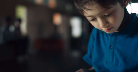 Close up curious focused schoolboy using tablet surfing internet in school hall. Obsessed with gadget teen boy playing online game alone. Elementary age guy ignore friends having fun on break.の写真素材