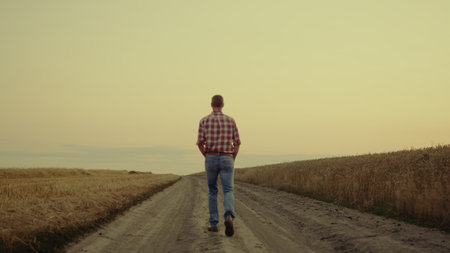Farmer man walking road alone at sunset field. Small business owner rancher agronomist examining wheat farm outdoor. Calm relax male agronom pass golden countryside landscape. Meditate nature conceptの写真素材