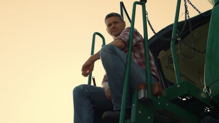 Successful farmer resting tractor at sunset. Tired agronomist inspecting crop field from harvester...
