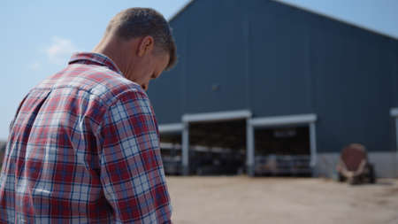 Farmer resting sitting agrocomplex yard after work on modern livestock facility close up. Agronomic specialist leaning on fence near large cow farm building. Agrobusiness owner relax outside cowshed.の写真素材