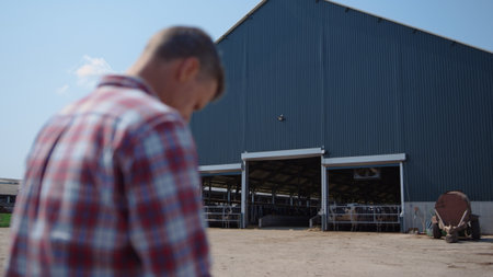 Farm worker sitting outside big barn facility on farmland ranch close up. Professional farmer resting after hard work feeding animals in modern countryside cowshed. Exterior of agricultural complex.の写真素材