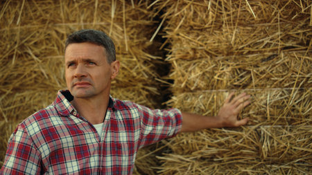 Portrait farmer inspect field in autumn. Focused farmland worker lean hay stack resting after working day. Thoughtful man agronomist observing seasonal harvesting. Agribusiness specialist concept.の写真素材