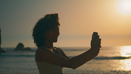 Pretty african american girl smiling on phone camera standing seashore at sunrise close up. Attractive happy woman making selfie on nature summer evening. Carefree lady taking photo on smartphone.の写真素材