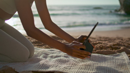 Unknown slim girl putting tablet on yoga mat lying sand beach close up. Young african american woman...