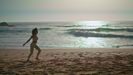 Energetic woman jogging on sandy beach enjoying beautiful sunrise. Happy slim girl wearing...