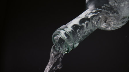 Fresh water splashing from glass bottle in super slow motion close up. Mesmerizing view of clear liquid stream flowing from transparent container on black background. Drink gurgling falling down.の写真素材