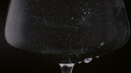 Small bubbles swirling inside full goblet of water in super slow motion close up. Crystal wineglass filled with clear liquid on black background. Drops of mineral drink falling from glass surface.の写真素材