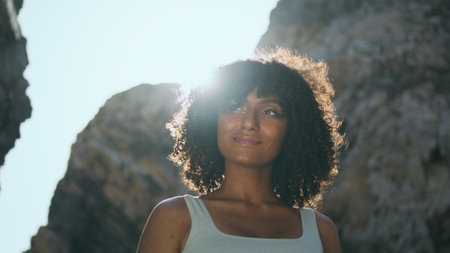 Smiling african american woman standing rocky beach lighted bright sunlight closeup. Portrait of curly young woman relaxing after workout on nature summer day. Calm sportswoman looking camera outdoorsの写真素材