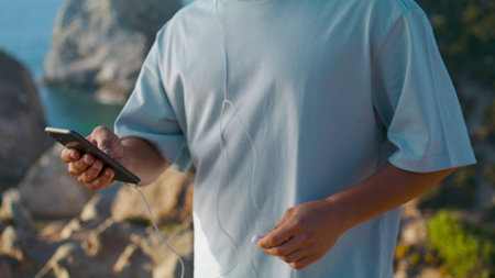 Man hands using smartphone at ocean edge cliff closeup. Athlete guy check phone holding earphones outdoors. Unrecognized user scrolling social media select podcast at sea. Smart technology concept.の写真素材