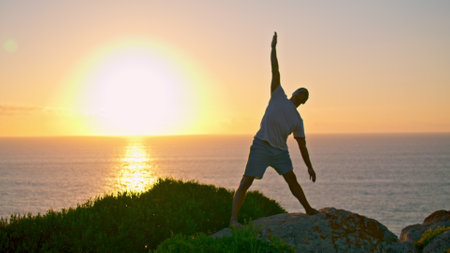 Muscular athlete silhouette stretching at sea rising sun. Strong man exercising on rock ocean edge. Unrecognized yoga guy relaxing enjoying sunset sunlight alone. Active sport lifestyle concept.の写真素材