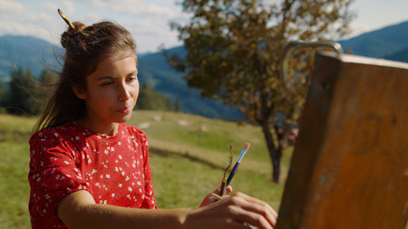 Closeup of inspired woman painter drawing picture in summer mountains. Portrait of talented girl creating artwork on green meadow. Focused artist painting using easel with brush in hair outdoors.の写真素材