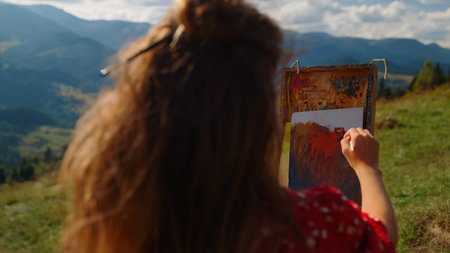 Back view of woman painter drawing with paintbrush beautiful mountains landscape close up. Unknown girl mixing paints on color palette standing green meadow. Young artist creating picture summer day.の写真素材