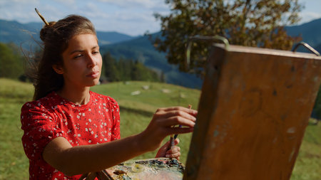Portrait of focused girl painter making brush stroke on easel summer day outdoors. Creative woman painting picture creating sketch of meadow closeup. Talented artist using color palette in mountainsの写真素材