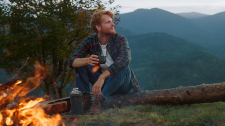 Travel man rest campfire on nature. Close up carefree guy enjoy mountains evening outside. Red hair man drink tea on vacation tourism activity. Relaxed hiker camping in forest. Tourist holiday conceptの写真素材