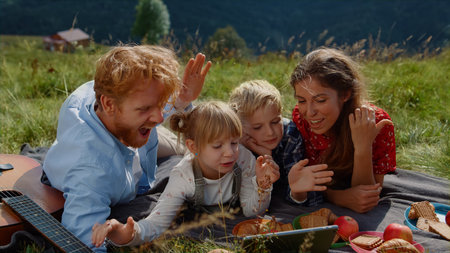 Cheerful family speaking online using tablet computer in mountains close up. Happy couple with children having video call on modern gadget outdoors. Joyful parents with kids waving hands to tab cameraの写真素材