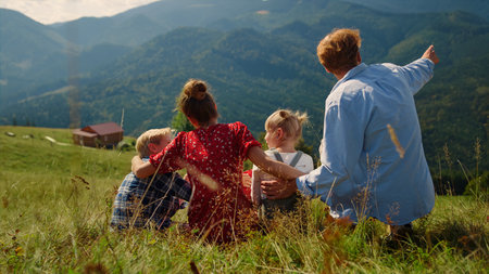 Happy parents sitting green grass hill with carefree children sunny day. Back view of cheerful family hugging on mountains meadow. Unrecognizable woman waving hands pointing finger on beautiful natureの写真素材
