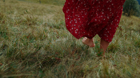 Unknown barefoot girl walking on green grass cloudy day close up. Bare woman feet making steps on meadow summer day. Unrecognizable young lady in red dress enjoying vacation in beautiful mountains.の写真素材