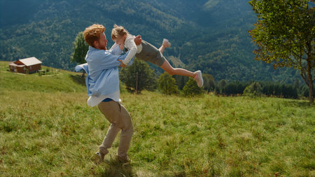 Playful ginger father circling daughter in arms sunny day standing green grass mountain meadow. Cheerful man playing with little lady on beautiful meadow. Happy guy spending summer vacation with childの写真素材