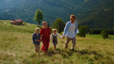 Positive family stepping green grass on walk to top green slope sunny day. Happy parents with two...