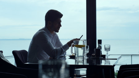 Handsome man scrolling smartphone touchscreen in modern restaurant. Thoughtful silhouette freelancer...