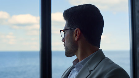Frustrated guy looking window considering business at sea panorama view closeup. Confident man ceo in glasses thinking relax in office. Successful bearded businessman contemplate investment planningの写真素材