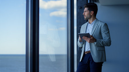 Happy businessman using tablet computer in ocean view office. Ceo professional reading good news in online app pad. Successful entrepreneur looking panoramic window. Corporate worker lifestyleの写真素材