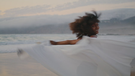 Cheerful excited girl spinning on gloomy beach with white shawl summer twilight. Curly african american woman waving pareo smiling at camera wearing black swimsuit. Happy brunette relaxing on nature.の写真素材