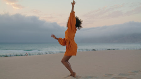 Emotional girl contemporary dancer waving hands performing on sand seashore summer evening. Smiling flexible african american woman wearing orange dress dancing modern style on gloomy beach.の写真素材