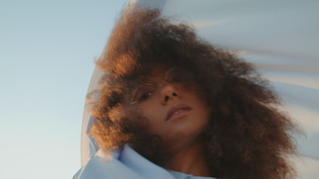 Sensual african american girl dancing with white fluttering fabric in front summer sky close up. Attractive curly woman performing on nature holding light cloth. Seductive brunette model posing cameraの写真素材