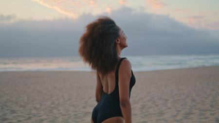 Relaxed woman tourist walking sandy gloomy beach in black swimsuit at summer dusk. Carefree african american girl relaxing on beautiful seashore at twilight. Attractive young brunette going near sea.の写真素材