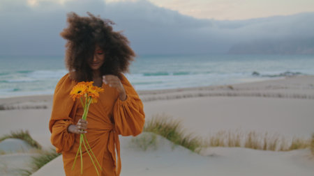 African american cheerful girl posing on windy gloomy beach with beautiful orange bouquet close up. Attractive young woman with curly hairstyle smiling camera standing on sand seashore with flowers.の写真素材