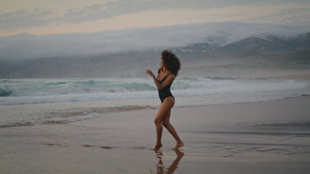 Beautiful sensual girl dancer performing graceful movements on wet beach summer evening. Attractive african american woman moving body smoothly dancing contemporary style. Brunette relax on seashore.の写真素材