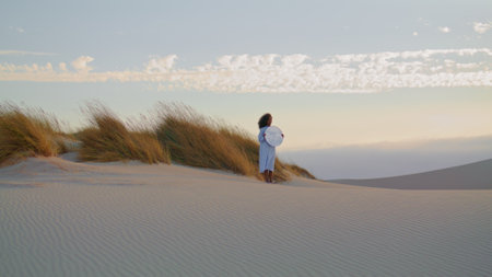 Unknown young woman with mirror standing on sand desert summer evening. African american girl posing in front gray sky with light clouds. Brunette wearing white dress holding speculum in hands.の写真素材