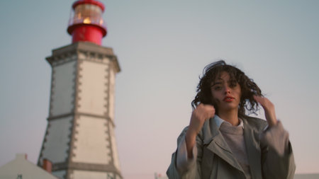 Beautiful girl resting lighthouse view. Casual young woman looking camera adjust curly hair. Worried pensive female contemplating walking seaside. Serious traveler turning enjoying evening trip aloneの写真素材