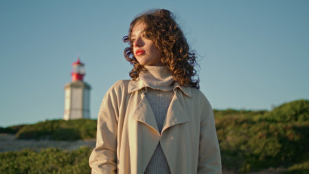Calm girl watching sunset at lighthouse alone. Curly pensive woman look camera relaxing quiet evening on green island. Thoughtful stylish female dream admiring nature in sunlight. Serene peaceful moodの写真素材