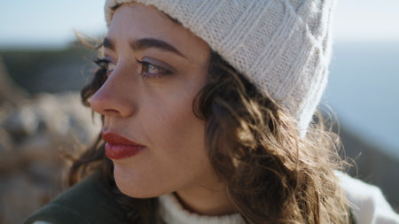 Closeup smiling girl outdoors rest on cold spring day. Cheerful traveler posing in knitted hat with curls swaying in wind. Beautiful young woman look camera enjoy ocean holidays. Serene happy female.の写真素材