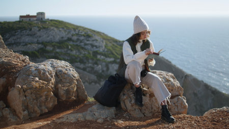 Romantic girl reading book on rocky mountain. Serene tourist resting ocean cliff enjoying tranquil...