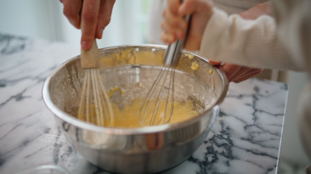 Baby woman hands mixing ingredients at bowl indoors closeup. Unknown family cooking pastry together at light kitchen. Anonymous child girl holding whisk helping mom. Mother fingers combining doughの写真素材