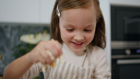 Laughing kid making dough at home portrait. Little smiling girl playing with pastry indoors close up. Funny child in apron having fun baking at cozy place. Positive baby helping mother in cooking roomの写真素材