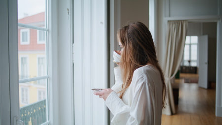 Calm woman sipping coffee at flat interior closeup. Thoughtful girl watching window holding saucer alone at domestic apartment. Smiling young lady looking camera posing at morning light portraitの写真素材