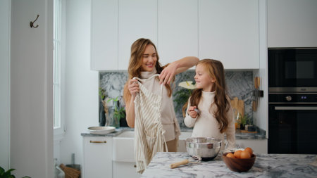 Cheerful woman putting apron on daughter light kitchen. Friendly family preparing cooking in apartment together. Single mother and baby girl spending time at home. Child having fun with female parentの写真素材