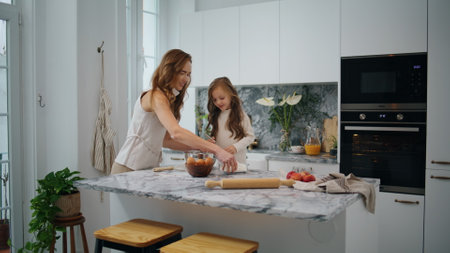 Cute family cooking at kitchen together. Young woman and child girl preparing dough on table. Mother daughter sifting flour in modern interior house. Adorable kid helping parent at light white homeの写真素材
