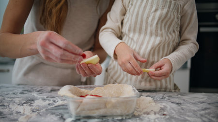 Woman child hands putting apple at baking form close up. Unrecognizable family preparing pie together at light kitchen. Unknown helping mother decorating future dessert together with daughterの写真素材